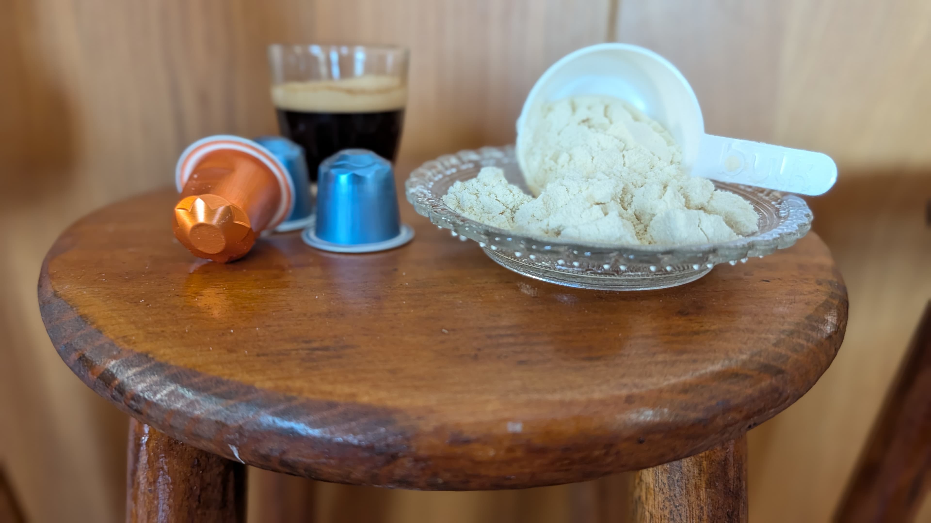 Creatina en polvo junto con vaso de café y cápsulas nespresso en taburete de madera